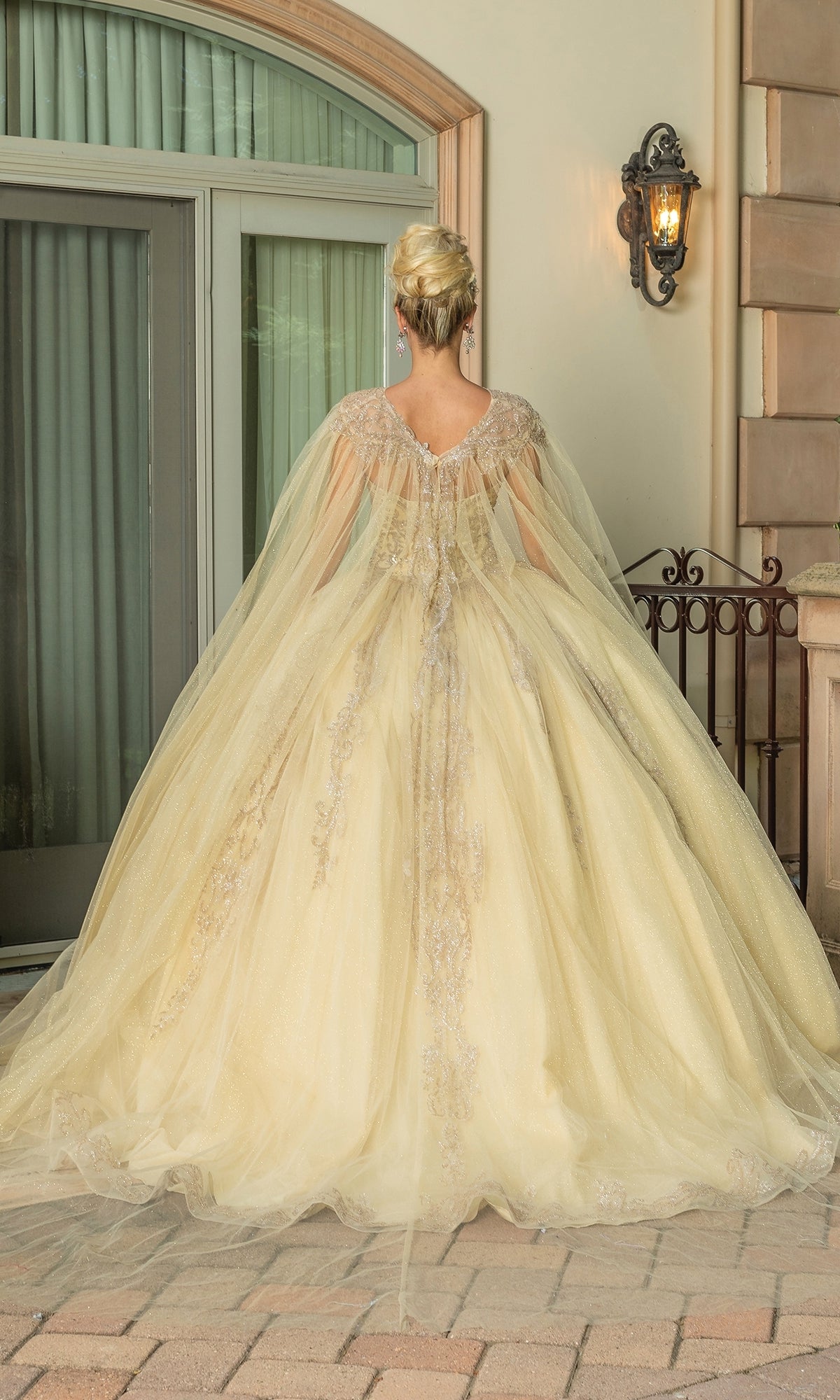A person in the Traditional Quinceañera Dress: Dancing Queen 1672, featuring light yellow fabric, ornate details, a long flowing skirt, and sheer cape, stands facing away before a large window and decorative wall sconce.